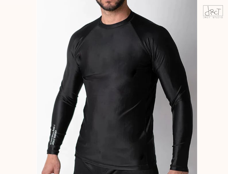 May include: A solid black long-sleeve compression shirt with a crew neckline and raglan sleeves. The fabric has a smooth, slightly reflective finish. A small white text graphic is printed vertically on the lower portion of the right sleeve.