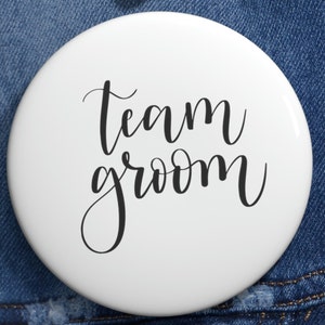 Op de afbeelding: Ronde witte knop met zwarte kalligrafie met de tekst "Team Groom".