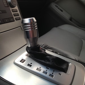 May include: Close-up of a silver car's gear shifter with a black leather gaiter. The shifter has a silver knob with a textured design and a silver plate with the letters "P", "R", "N", and "D" for the gear positions. The shifter also has a button labelled "Shift Lock".