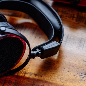 May include: Black headphones with a red wood veneer on the earcups. The headphones are on a wooden surface.