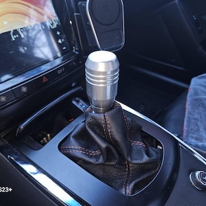 May include: Close-up of a silver gear shift knob with a black leather boot. The knob has a textured design and is installed in a car.