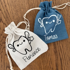 May include: Two small, personalised tooth fairy bags. One is white with the name Florence and a black and white tooth fairy illustration. The other is blue with the name Tomas and a white tooth fairy illustration.