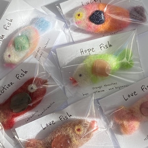May include: A collection of needle-felted fish ornaments in clear plastic packaging, each featuring a unique gemstone accent. Labels include Lucky Fish, Hope Fish, Love Fish, and others, with text detailing the stone and theme.