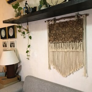 May include: A woven wall hanging with a natural, earthy aesthetic. The upper portion features a textured, brown material, while the lower section showcases cream-colored macrame fringe. The piece is suspended from a wooden rod, adding a rustic touch to the decor.
