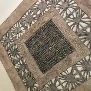 May include: A square-shaped wall hanging made of woven paper with a geometric design. The paper is a light brown color and the design is a darker brown color.