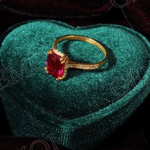 Op de afbeelding: Een gouden ring met een grote, vierkante rode edelsteen omringd door kleinere witte edelstenen. De ring wordt gepresenteerd in een groene fluwelen hartvormige doos.