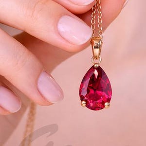 May include: A gold-colored necklace with a teardrop-shaped, deep red gemstone pendant. The pendant is held by a gold setting and hangs from a delicate gold chain. The word "Gemoj" is faintly visible in the image.
