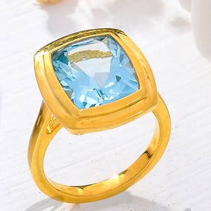 May include: A gold ring featuring a square, light blue gemstone. The ring has a simple band and a bezel setting. The word "Gemoj" is written in cursive on the surface. The ring is displayed on a white surface with dried flowers.