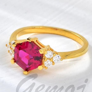 May include: A gold ring featuring a large, faceted, magenta gemstone at the center, surrounded by smaller white gemstones. The ring has a simple band and is set against a white background. The word "Gemoj" is written in cursive.