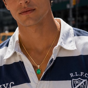 May include: A long-sleeve rugby shirt featuring wide navy blue and white horizontal stripes, a white collar, and a button placket. The shirt displays the text NYC RUGBY and a crest with R.L.F.C. 1967 167. A gold chain necklace with a green emerald pendant.