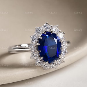 May include: A silver ring with a large, oval, deep blue gemstone encircled by smaller, clear stones. The ring's polished band highlights the central stone. The design is elegant and eye-catching.
