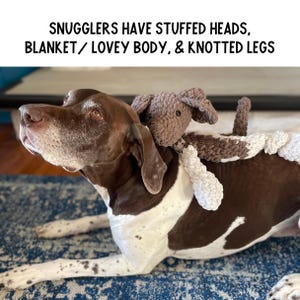 May include: A brown and white dog with a brown and white crocheted stuffed animal toy on its back. The toy has a knotted body and legs. The dog is lying on a blue and white patterned rug. The text "Snugglers have stuffed heads, blanket/lovey body, & knotted legs" is above the image.