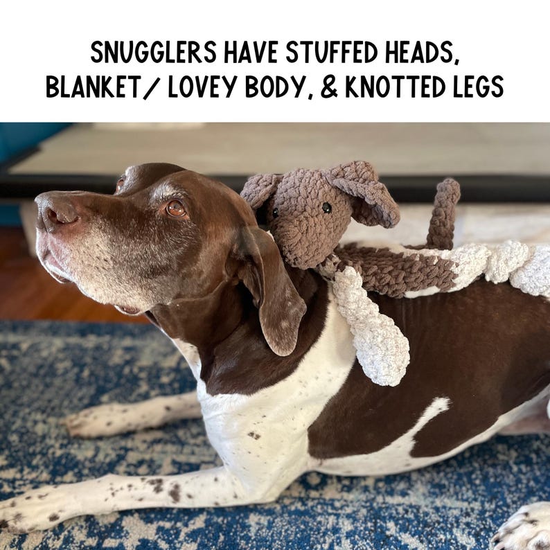 May include: A brown and white dog with a brown and white crocheted stuffed animal toy on its back. The toy has a knotted body and legs. The dog is lying on a blue and white patterned rug. The text "Snugglers have stuffed heads, blanket/lovey body, & knotted legs" is above the image.