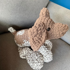 May include: A handmade plush dog toy in shades of brown and white. The dog has floppy ears, a small tail, and a black button eye. The toy is made of a soft, textured material, and is sitting on a gray surface.