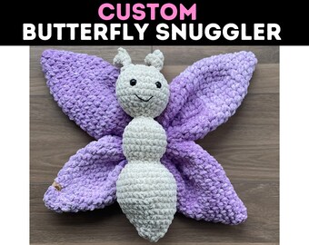 May include: A crocheted butterfly snuggle toy with a purple and white color scheme. The butterfly has a smiling face and a soft, plush texture.