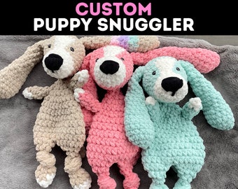 May include: Three crocheted puppy snugglers in beige, pink, and light blue. Each plush toy has a white face with black eyes and a black nose.
