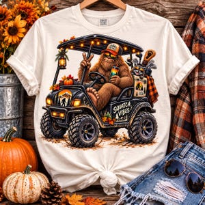 May include: A cream-colored t-shirt featuring a graphic of a brown Bigfoot driving a black golf cart. The Bigfoot wears a hat and sunglasses, holding a drink and making a peace sign. The text "SQUATCH VIBES" is printed on the side of the cart.