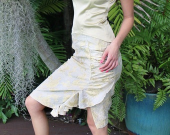 May include: A woman wearing a light green sleeveless top and a matching skirt with a ruffle hem. She is standing in front of a tropical plant.