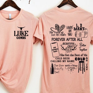 May include: Two light pink t-shirts hanging on wooden hangers. The front of the first shirt has the text "LUKE COMBS" with a bull graphic. The back of the second shirt has song lyrics and graphics in black.