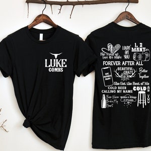 May include: Two black t-shirts with white text. One shirt features "LUKE COMBS" with a bull skull logo. The other shirt has song lyrics and phrases like "Forever After All" and "Cold Beer Calling My Name".