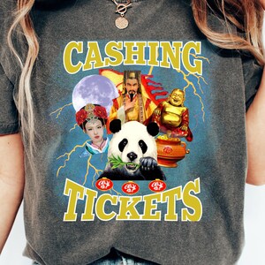 May include: Dark gray t-shirt with a graphic featuring a panda eating bamboo, a woman, a man in gold robes, and a laughing Buddha. The text "CASHING TICKETS" is in yellow.