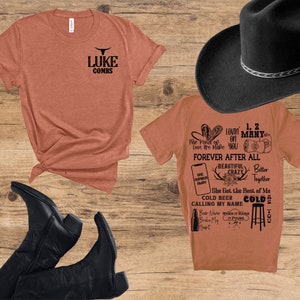 May include: An assortment of country-themed items on a wooden surface. Includes two orange t-shirts with black text, a black cowboy hat, and black cowboy boots. One shirt features the text "LUKE COMBS" and a bull graphic. The other shirt has various phrases.