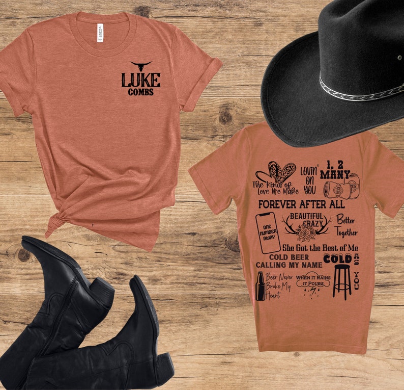 May include: An assortment of country-themed items on a wooden surface. Includes two orange t-shirts with black text, a black cowboy hat, and black cowboy boots. One shirt features the text "LUKE COMBS" and a bull graphic. The other shirt has various phrases.