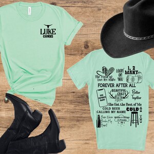 May include: Two mint green t-shirts with black text and a black cowboy hat are displayed on a wooden surface. One shirt reads "LUKE COMBS" and the other has song lyrics. Black cowboy boots are also visible.