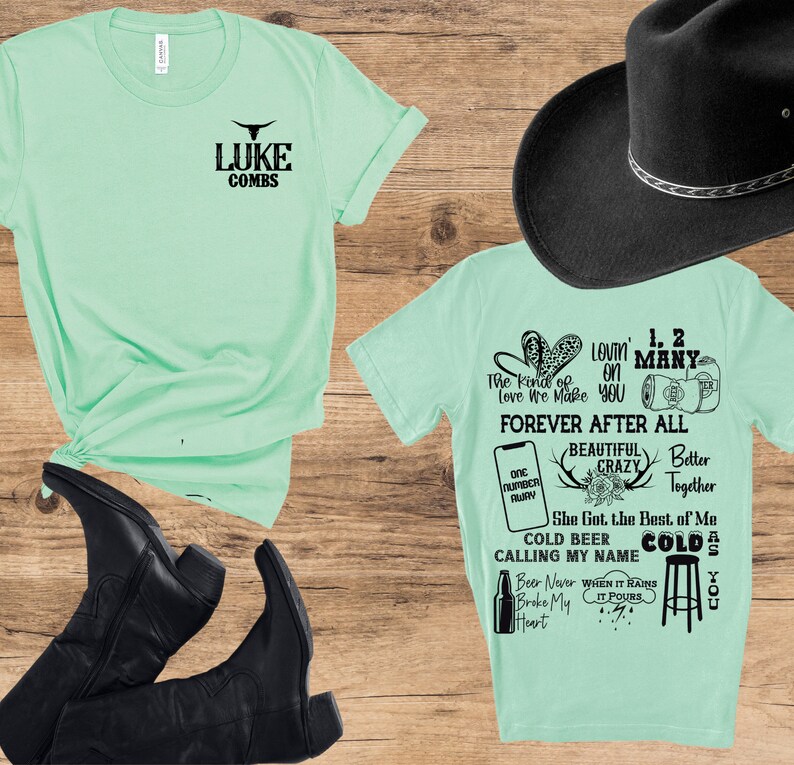 May include: Two mint green t-shirts with black text and a black cowboy hat are displayed on a wooden surface. One shirt reads "LUKE COMBS" and the other has song lyrics. Black cowboy boots are also visible.