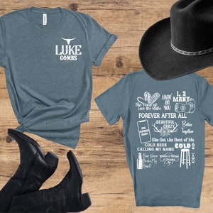 May include: Two blue t-shirts with white text and a black cowboy hat. One shirt says "LUKE COMBS" with a bull skull logo. The other shirt has song lyrics and phrases like "Forever After All" and "Cold Beer". Black cowboy boots are also visible.