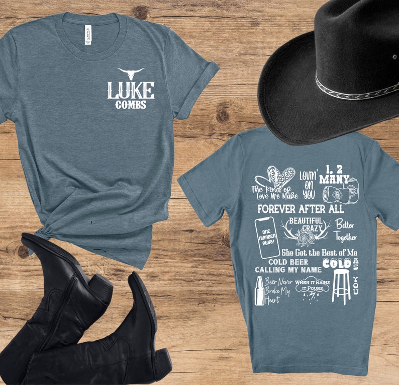 May include: Two blue t-shirts with white text and a black cowboy hat. One shirt says "LUKE COMBS" with a bull skull logo. The other shirt has song lyrics and phrases like "Forever After All" and "Cold Beer". Black cowboy boots are also visible.