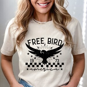 May include: A beige t-shirt with a black graphic of an eagle with the text "FREE BIRD" above it and "1973 america" below it.