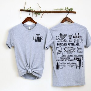 May include: Two gray t-shirts on wooden hangers. The front of one shirt has the text "LUKE COMBS" with a bull graphic. The back of the other shirt features various graphics and text, including "FOREVER AFTER ALL" and "COLD BEER CALLING MY NAME".
