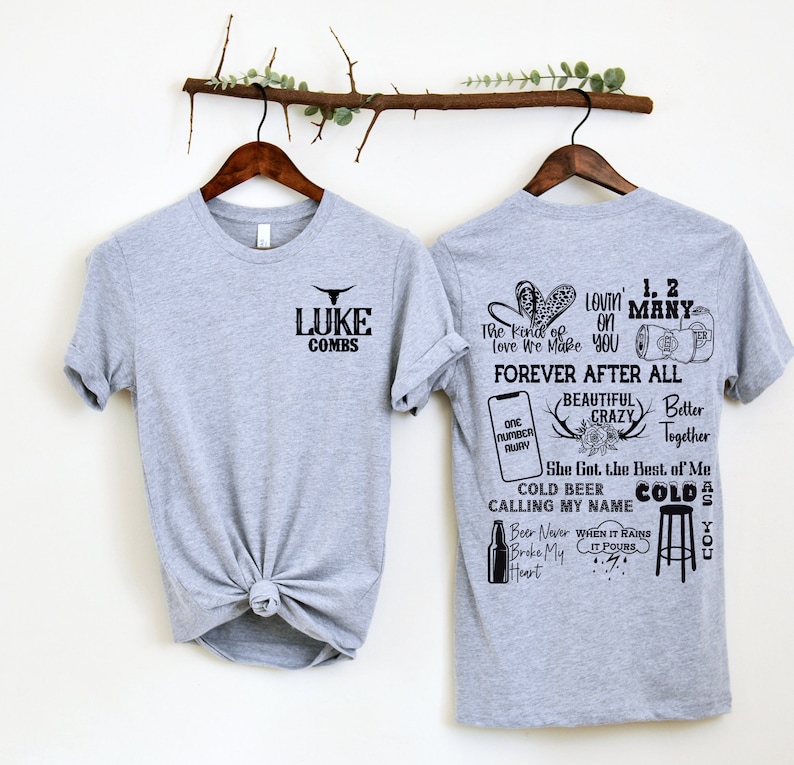 May include: Two gray t-shirts on wooden hangers. The front of one shirt has the text "LUKE COMBS" with a bull graphic. The back of the other shirt features various graphics and text, including "FOREVER AFTER ALL" and "COLD BEER CALLING MY NAME".