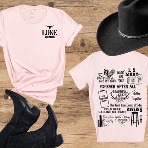 May include: A light pink t-shirt with the text "LUKE COMBS" and a bull graphic. A second light pink t-shirt features song lyrics and graphics. Black cowboy boots and a black cowboy hat are also in the image.