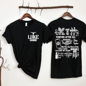 May include: Two black t-shirts on wooden hangers. The front of one shirt has "LUKE COMBS" and a bull graphic in white. The back of the other shirt has song lyrics and graphics in white, including "FOREVER AFTER ALL" and "COLD BEER".