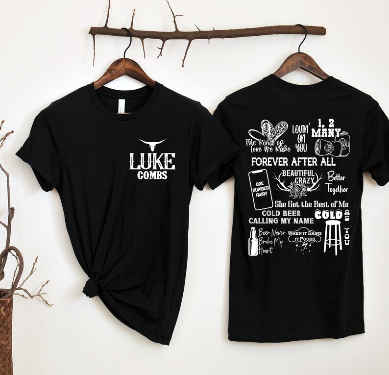 May include: Two black t-shirts on wooden hangers. The front of one shirt has "LUKE COMBS" and a bull graphic in white. The back of the other shirt has song lyrics and graphics in white, including "FOREVER AFTER ALL" and "COLD BEER".