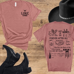 May include: A dusty rose t-shirt with the text "LUKE COMBS" and a bull skull logo. The back of the shirt features song lyrics and phrases like "Forever After All" and "Cold Beer Calling My Name". A black cowboy hat and boots are also in the image.