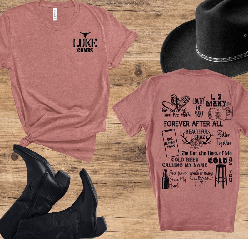 May include: A dusty rose t-shirt with the text "LUKE COMBS" and a bull skull logo. The back of the shirt features song lyrics and phrases like "Forever After All" and "Cold Beer Calling My Name". A black cowboy hat and boots are also in the image.