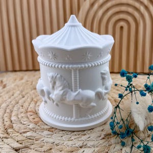 Può includere: Una scatola decorativa bianca a tema giostra. La scatola presenta un design dettagliato di cavalli della giostra e una parte superiore a forma di tenda. La scatola è realizzata in un materiale solido ed è adatta per riporre piccoli oggetti.