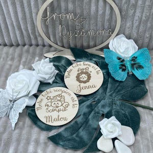 May include: Two wooden plaques with engraved text and illustrations. The first plaque is light brown and has the text 'Notre bébé lion est là' with a lion illustration and the name 'Jenna'. The second plaque is light brown and has the text 'Notre bébé scorpion est là' with a scorpion illustration and the name 'Malou'.