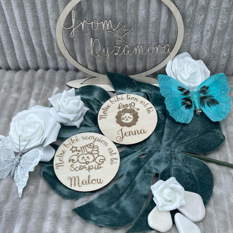 May include: Two wooden plaques with engraved text and illustrations. The first plaque is light brown and has the text 'Notre bébé lion est là' with a lion illustration and the name 'Jenna'. The second plaque is light brown and has the text 'Notre bébé scorpion est là' with a scorpion illustration and the name 'Malou'.