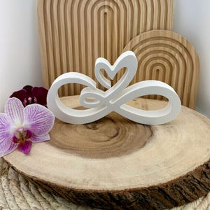 Peut inclure: Une sculpture blanche en forme de symbole infini avec un cœur, posée sur une tranche de bois avec une orchidée violette et blanche. Le fond présente des éléments décoratifs en bois arqués. Objet décoratif.