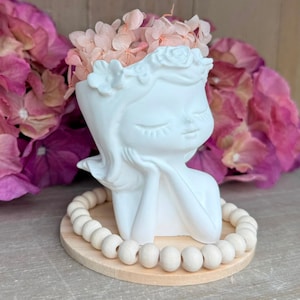Può includere: Un vaso in ceramica bianca a forma di testa con occhi chiusi e corona floreale, contenente petali di ortensia rosa chiaro. Il vaso poggia su una base circolare in legno circondata da una fila di perline di legno lisce e di colore chiaro.