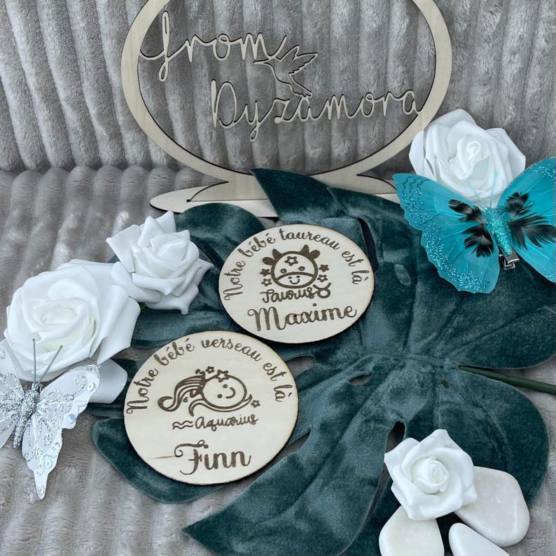 May include: Two wooden plaques with engraved text and images. The top plaque has a bull image and the text 'Notre bébé taureau est là Maxime Taurus'. The bottom plaque has a mermaid image and the text 'Notre bébé verseau est là Finn Aquarius'. Both plaques are on a teal green surface with white flowers and a silver butterfly.