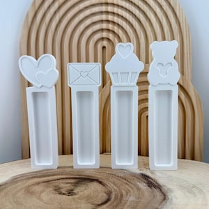May include: Four white silicone molds with different shapes: hearts, an envelope, a cupcake, and a teddy bear. Each mold has a rectangular base. The molds are arranged in a row against a wooden background with a rainbow design.