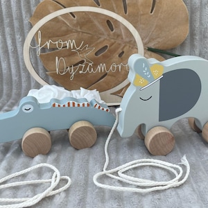 Op de afbeelding: Een set van twee houten trek speelgoed, een blauwe krokodil en een grijze olifant met een gele en witte gepatroneerde hoed. De speelgoedjes zijn verbonden door een wit touw.