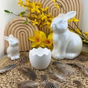 Puede incluir: Una composición decorativa de temática pascual con dos figuras de conejos de cerámica blanca, un recipiente blanco en forma de huevo y flores amarillas. La escena está dispuesta sobre una estera tejida con plumas dispersas.