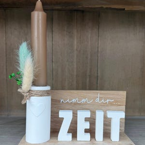Puede incluir: Un portavelas decorativo con una vela marrón, base blanca y elementos de madera. La base blanca tiene un diseño de corazón y está adornada con plumas y vegetación. El letrero de madera dice "nimm dir ZEIT".