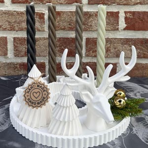 Puede incluir: Una bandeja decorativa blanca con una cabeza de ciervo y figuras de árboles de Navidad. Velas retorcidas en negro, marrón y verde se alzan detrás de la bandeja. Adornos dorados y vegetación añaden acentos festivos. Una etiqueta "Hand Made" está unida a un árbol.